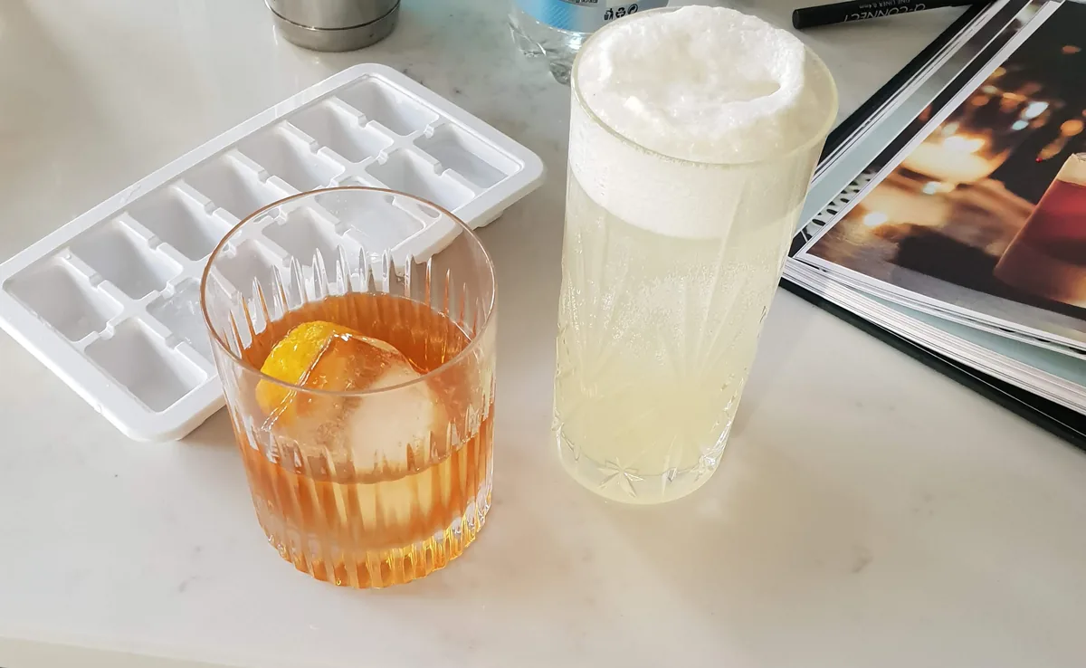Two homemade cocktails on a marble counter: an old fashioned with orange peel and a foam-topped gin fizz, with an ice cube tray and cocktail shaker behind